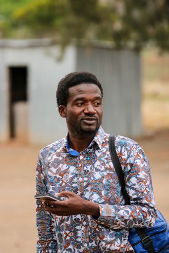 A young Black teacher in a colorful shirt with a phone on his hand arriving at school