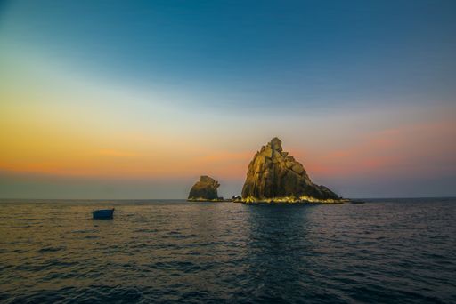 A scenic picture of a rocky islet deserted in ocean with a boat floating towards it during an amazing sunset