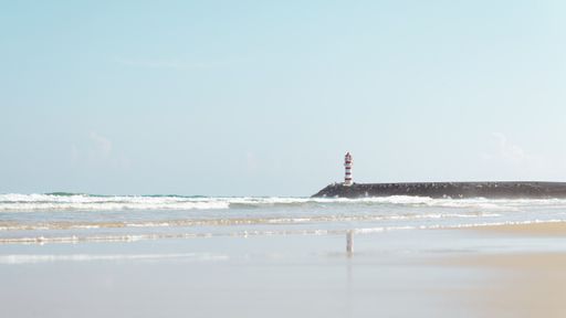 A view of a sandy beach with waves washing it and a light house nearby on a sunny day