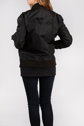 A young female wearing a black jacket standing against a white wall