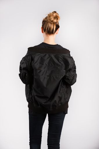 A young female wearing a black jacket standing against a white wall