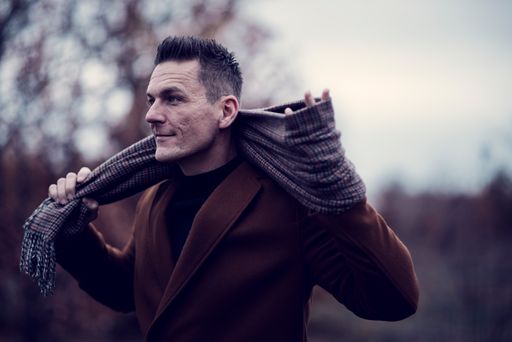 A portrait of a stylish Caucasian male with a brown blazer holding a gray scarf with blurred garden background