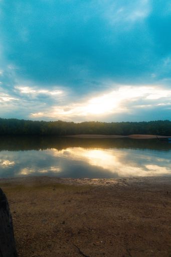 A vertical shot of a lake with sandy shore surrounded by trees in the distance during sunset