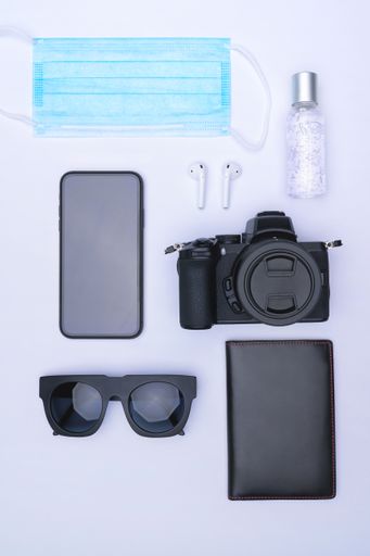 A vertical top view of a mask, hand sanitizer, ear pads, smartphone, camera, sunglasses and passport holder on a white background