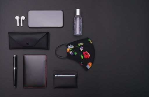 A top view of a smartphone, ear pads, hand sanitizer, mask, sunglasses case, ballpen, notebook, and cardholder on a black background