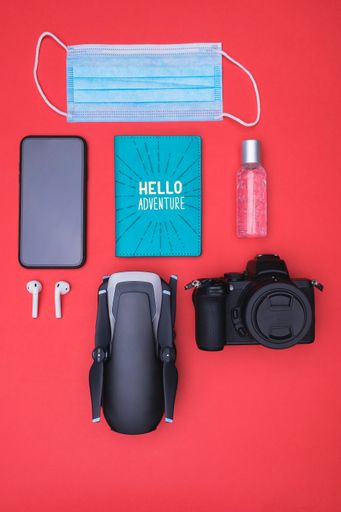 A vertical top view of a mask, hand sanitizer  smartphone, ear pads, passport holder, camera, and drone on a red background