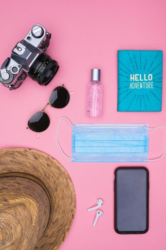 A top view of a smartphone, ear pads, mask, sunglasses, hand sanitizer, camera, passport holder, and hat on a pink background