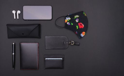 A top view of a smartphone, ear pads, luggage tag, mask, sunglasses case, ballpen, notebook, and cardholder on a black background