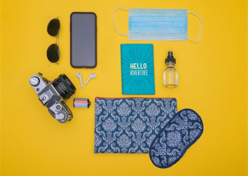 A top view shot of sunglasses, smartphone, ear pads, camera, film, passport holder, mask, sprayer, and pouch with a sleep mask on a yellow background