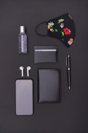 A vertical top view shot of a hand sanitizer, mask, cardholder, ear pads, smartphone, ballpen, and passport holder on a black background