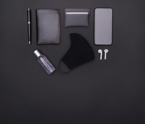 A top view of ballpen, passport holder, cardholder, smartphone, ear pads, mask, and hand sanitizer on a black background