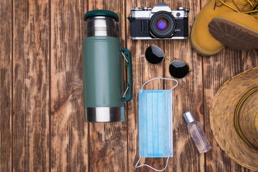 A top view shot of a camera, sunglasses, mask, hand sanitizer, shoes, hat, and tumbler on a wooden background