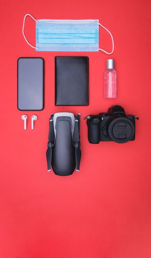 A vertical top view of a mask, hand sanitizer  smartphone, ear pads, passport holder, camera, and drone on a red background