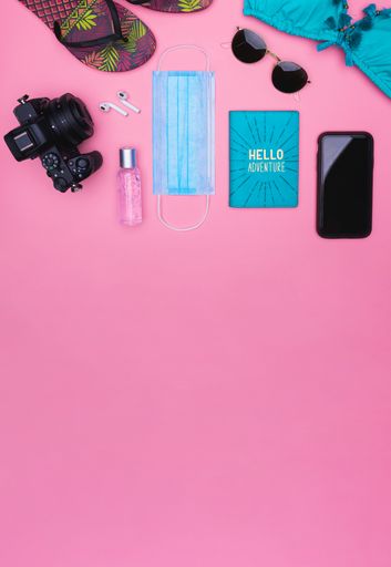 A vertical top view of a camera, ear pads, sanitizer, mask, passport holder, smartphone, sunglasses, bikini top and slippers on a pink background