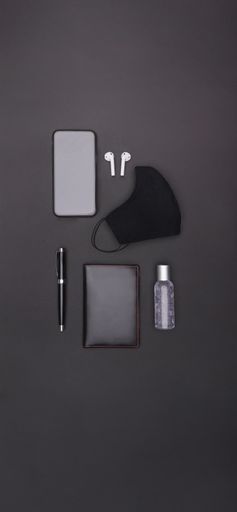 A vertical top view of a smartphone, ear pads, mask, ballpen, hand sanitizer and a passport holder on a black background