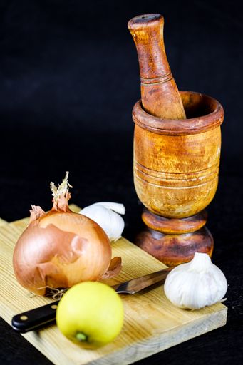 A vertical shot of a wooden mortar and pestle and garlic, onion and lemon on wooden chopping board on black background