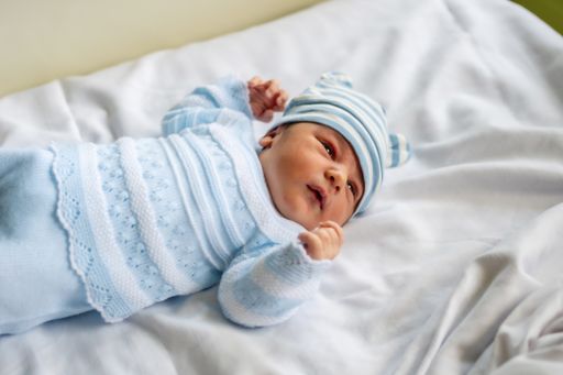 A photo of a newborn boy in the hospital, first day of life, with a white bodysuit, a blue hat and lying in the crib