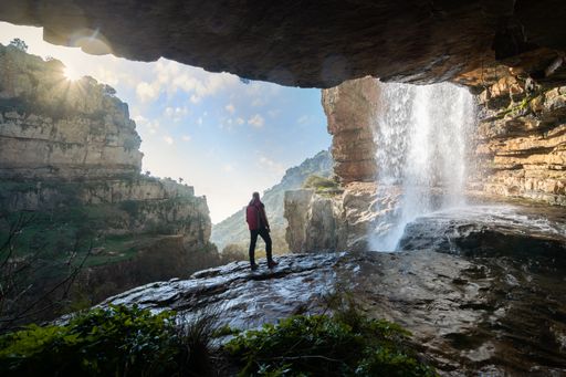 A beautiful shot of a male standing near a waterfall surrounded by cliffs under a blue sky