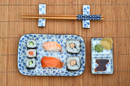 A top view high angle shot of sushi rolls, sauce, and dumplings in plates with floral patterns next to chopsticks on a bamboo table cover