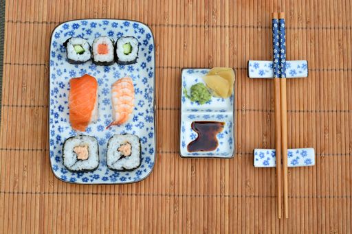 A top view high angle shot of sushi rolls, sauce, and dumplings in plates with floral patterns next to chopsticks on a bamboo table cover