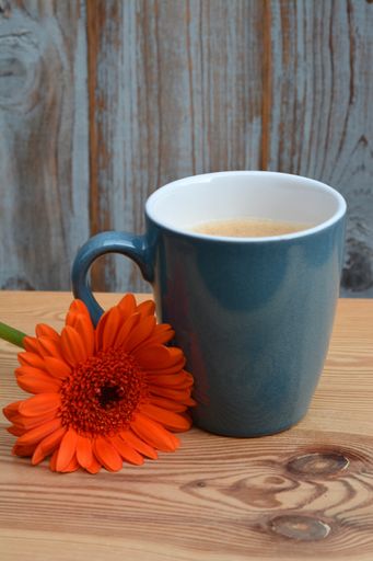 A vertical shallow focus shot of an orange Barberton Daisy flower lying next to a blue mug of coffee on a wooden table