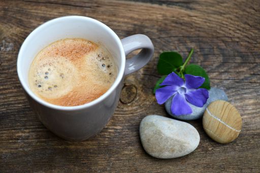 A high angle shot of coffee in a white mug next to pebbles and a Periwinkle flower on a wooden surface