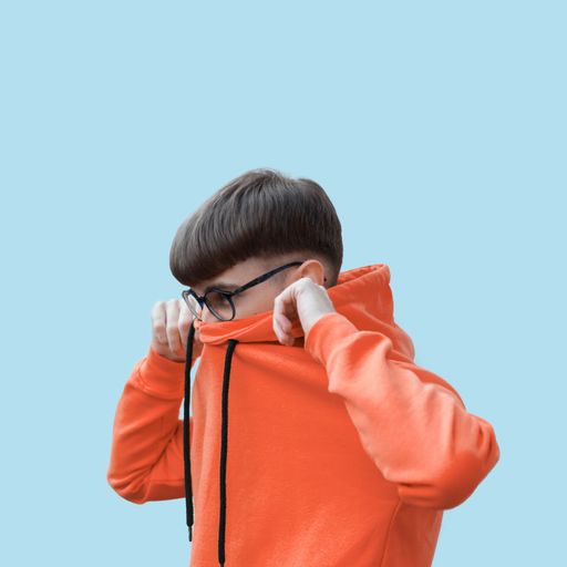 A closeup shot of a young male covering his face with an orange hoodie with glasses and a neat hairstyle on a light blue background