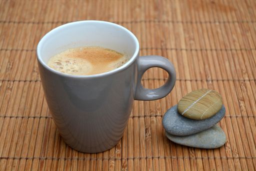 A high angle shallow focus shot of a white mug of coffee next to stacked pebbles on a bamboo table cover