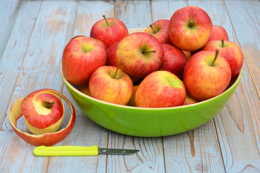 A closeup shot of red and yellow apples in a green plate next to a peeled apple on a wooden surface