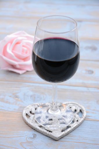 A vertical shallow focus shot of a red wine glass with a heart-shaped base with a pink rose on a wooden table