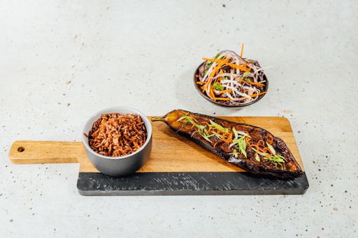 An overhead shot of a bowl of vegetables with red rice and eggplant on a wooden chopping board