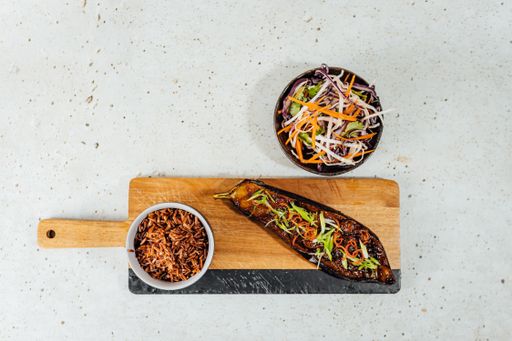 A top view of a bowl of vegetables with red rice and eggplant on a wooden chopping board
