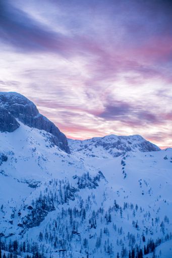 The vertical low angle shot of snowy mountain high in the winter with a pink-orange blurred skyline background