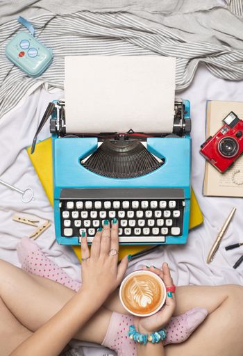 a vertical shot of girl with cofee typing on typewriter in bed