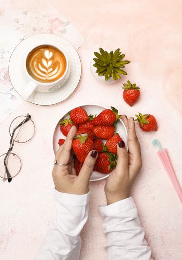 The female hands taking a strawberry from a plate by a cup of coffee and glasses