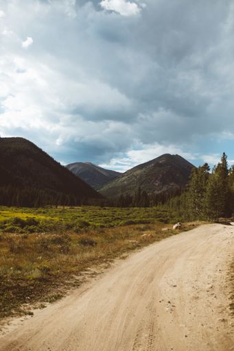 A vertical image of a dirt round lined with grasses and hills under a cloudy sky