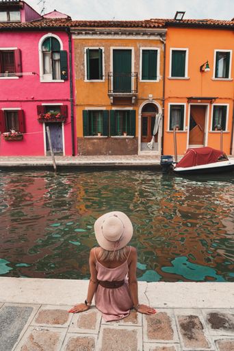 A vertical shot of a female model from behind sitting on the edge of a Grand canal against colorful houses of Burano, Venice