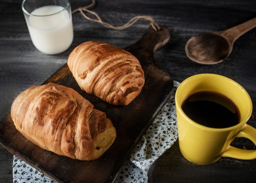 A closeup shot of the croissants on a wooden board along with a yellow mug of coffee, a wooden spoon and a cup of milk