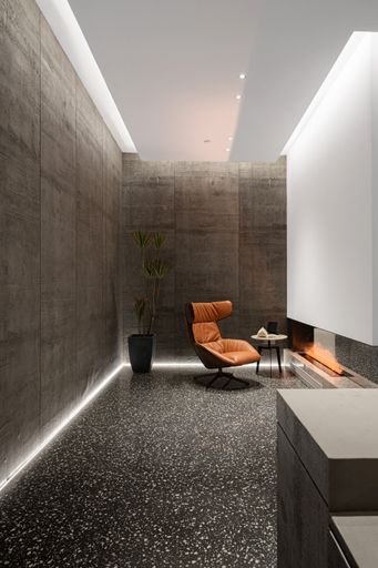 A vertical shot of a modern interior with an armchair, a small table, and artificial flowers next to a fireplace against the brown wall