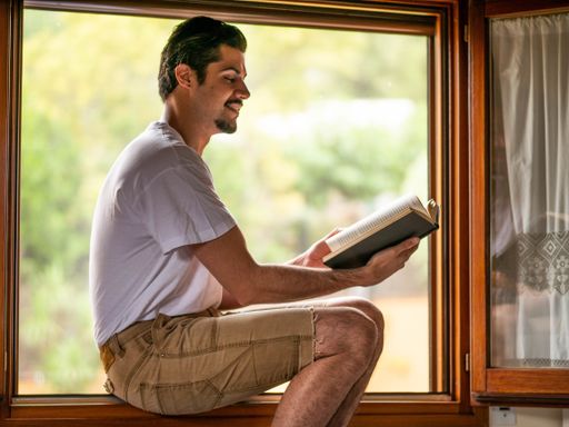 A picture of a brunette male sitting next to the window and reading a book