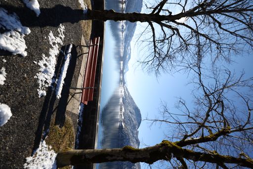 A vertical shot of a park bench against a lake reflecting mountains in winter