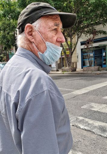 A vertical closeup of an old man waiting to cross the street, with a face mask and a cap.