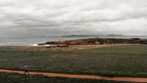 A wide shot of a person running on the pathway in the middle of grassy fields with a sea in the distance under a cloudy sky