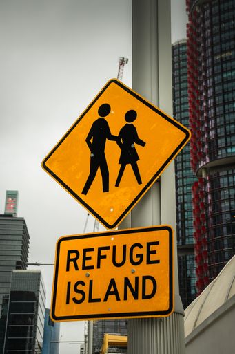 A vertical closeup shot of a yellow pedestrian street sign "Refuge Island" with a background of high buildings in Sydney, Australia