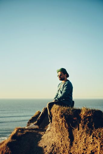 A vertical shot of a male with hats and sunglasses sitting on top of a hill against the seascape