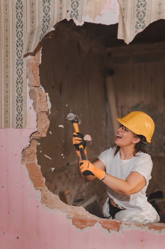 A professional Caucasian female construction worker wearing protective gear and smashing a hammer through the wall of an old house during renovation