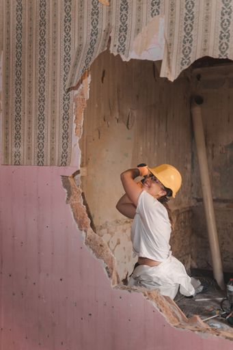 A professional Caucasian female construction worker wearing protective gear and smashing a hammer through the wall of an old house during renovation