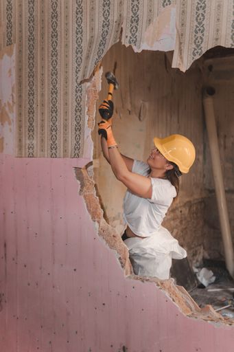 A professional Caucasian female construction worker wearing protective gear and smashing a hammer through the wall of an old house during renovation