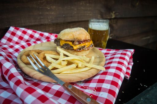 An angle shot of a burger on a wooden plate with onion rings and french fries next to a glass of beer.