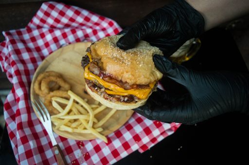 A close-up shot of hands holding a burger in the background of onion rings and french fries on a wooden plate.